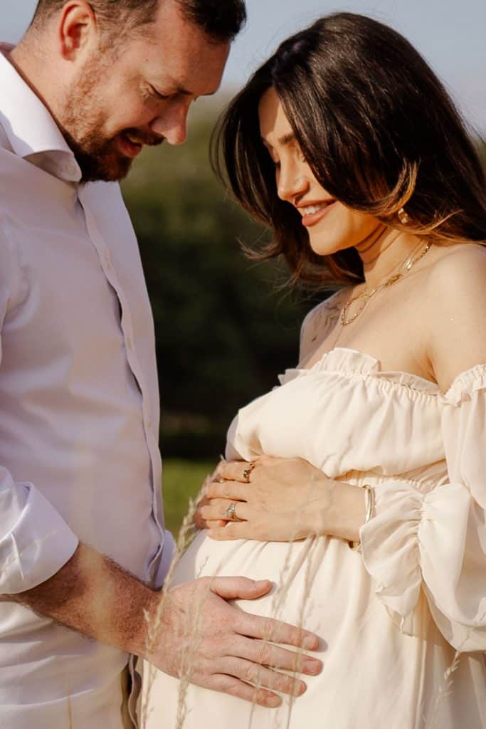 Pregnant couple standing together in a field with their hands resting on her bump