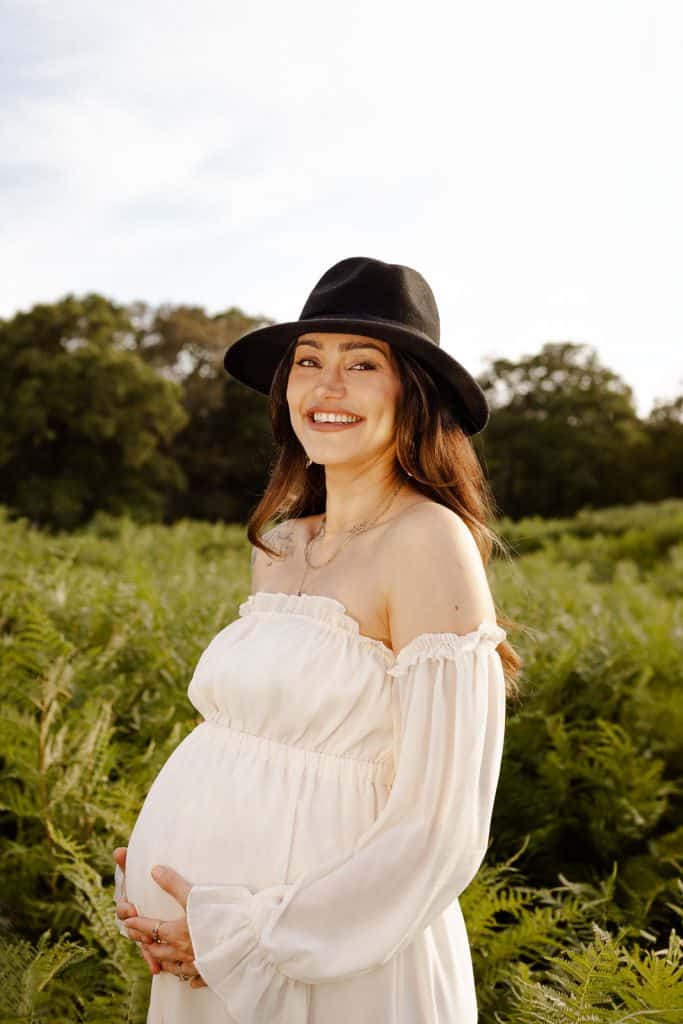 Pregnant woman in a white dress and black hat smiling during an outdoor maternity photography session