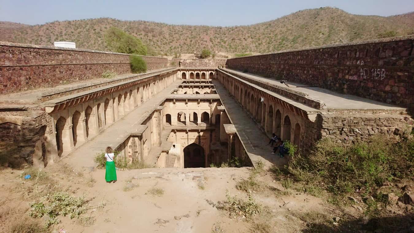 A long, deep stepwell lined with symmetrical rows of stone arches and descending levels, surrounded by dry hills in Rajasthan, India.