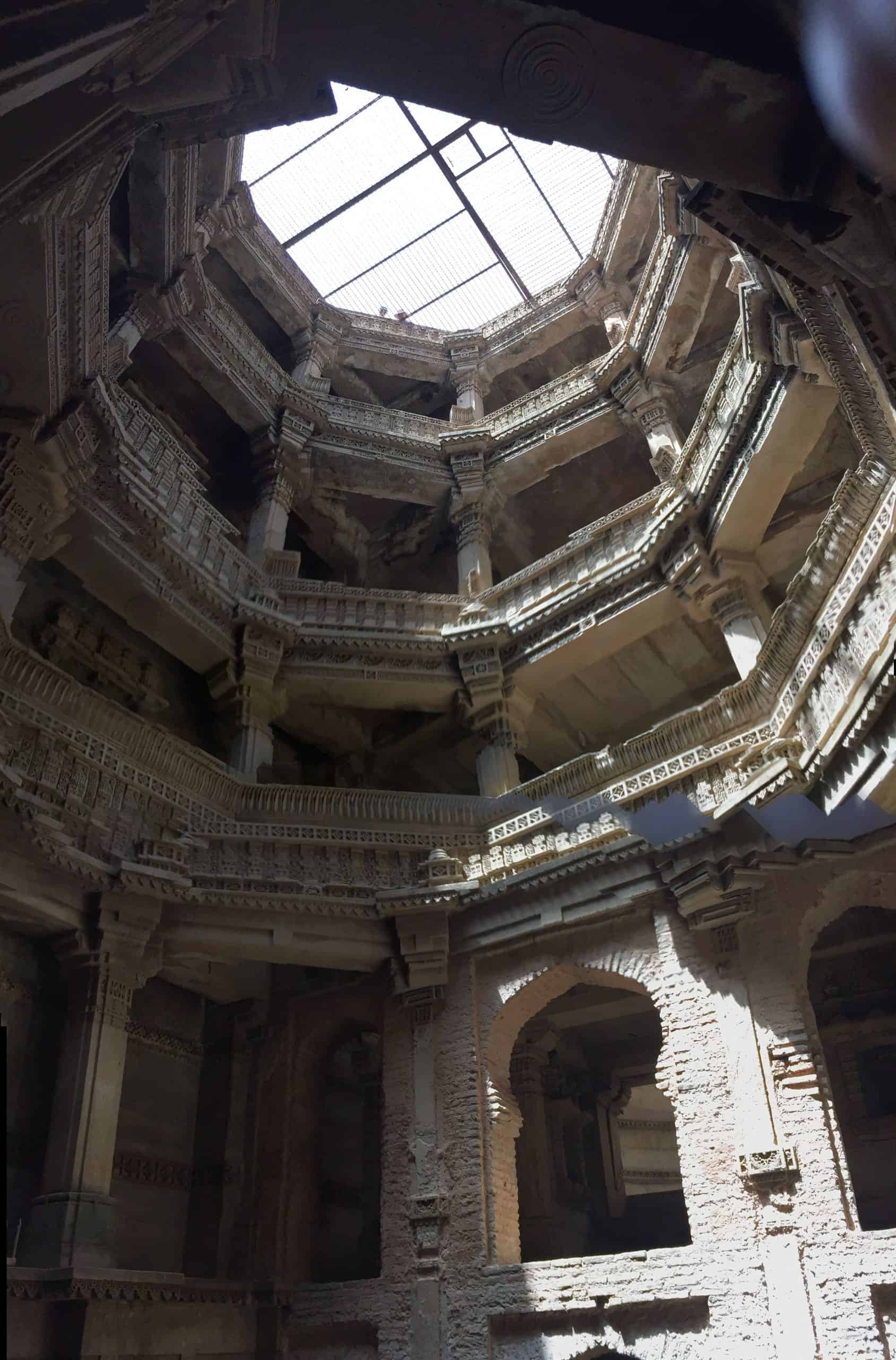 Ornate carved stone galleries rise through several levels of an Indian stepwell towards a bright, mesh-covered opening in the roof.