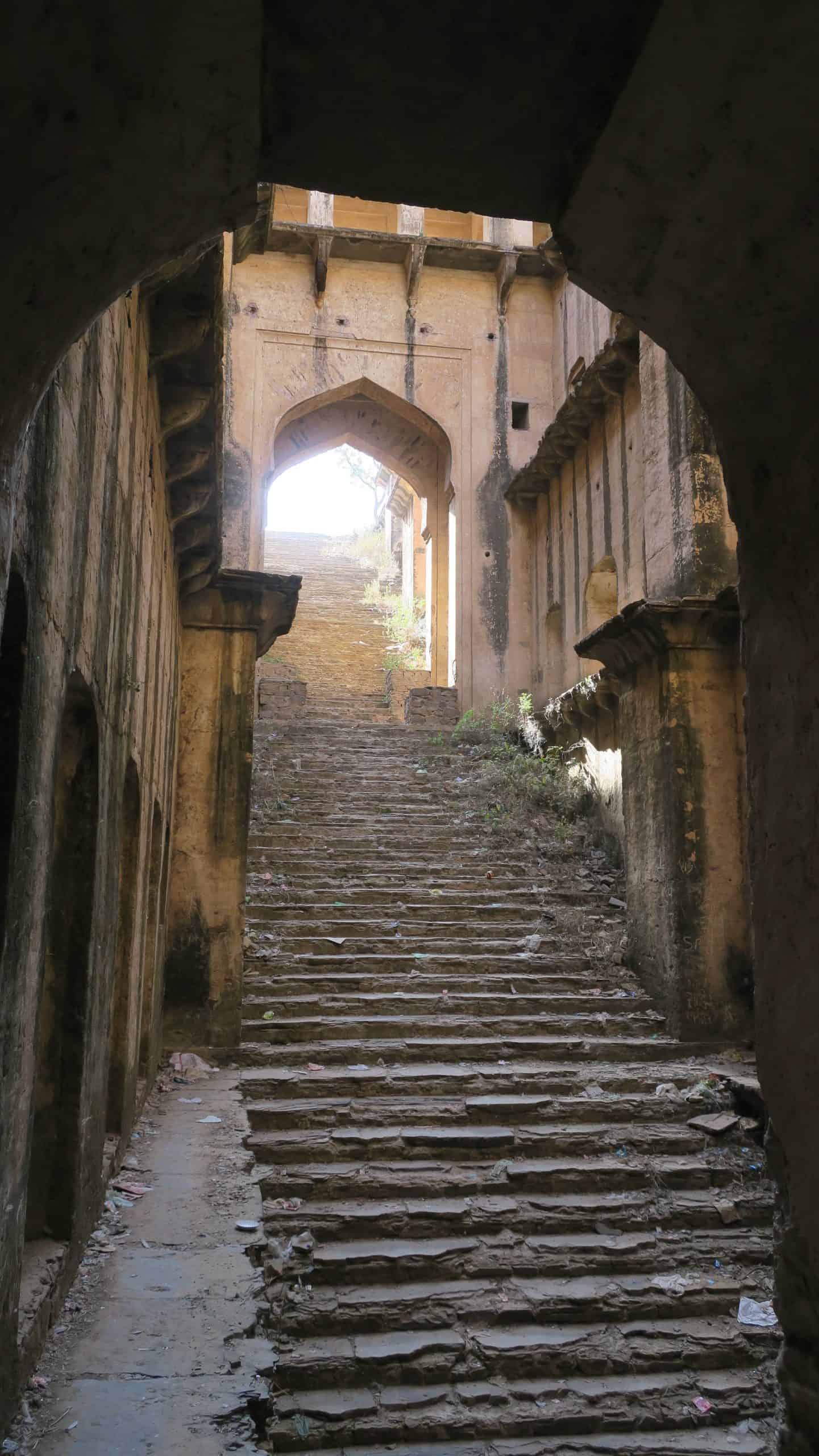 Worn stone steps climb through a shadowed arched passage towards the sunlight in an old Indian stepwell.