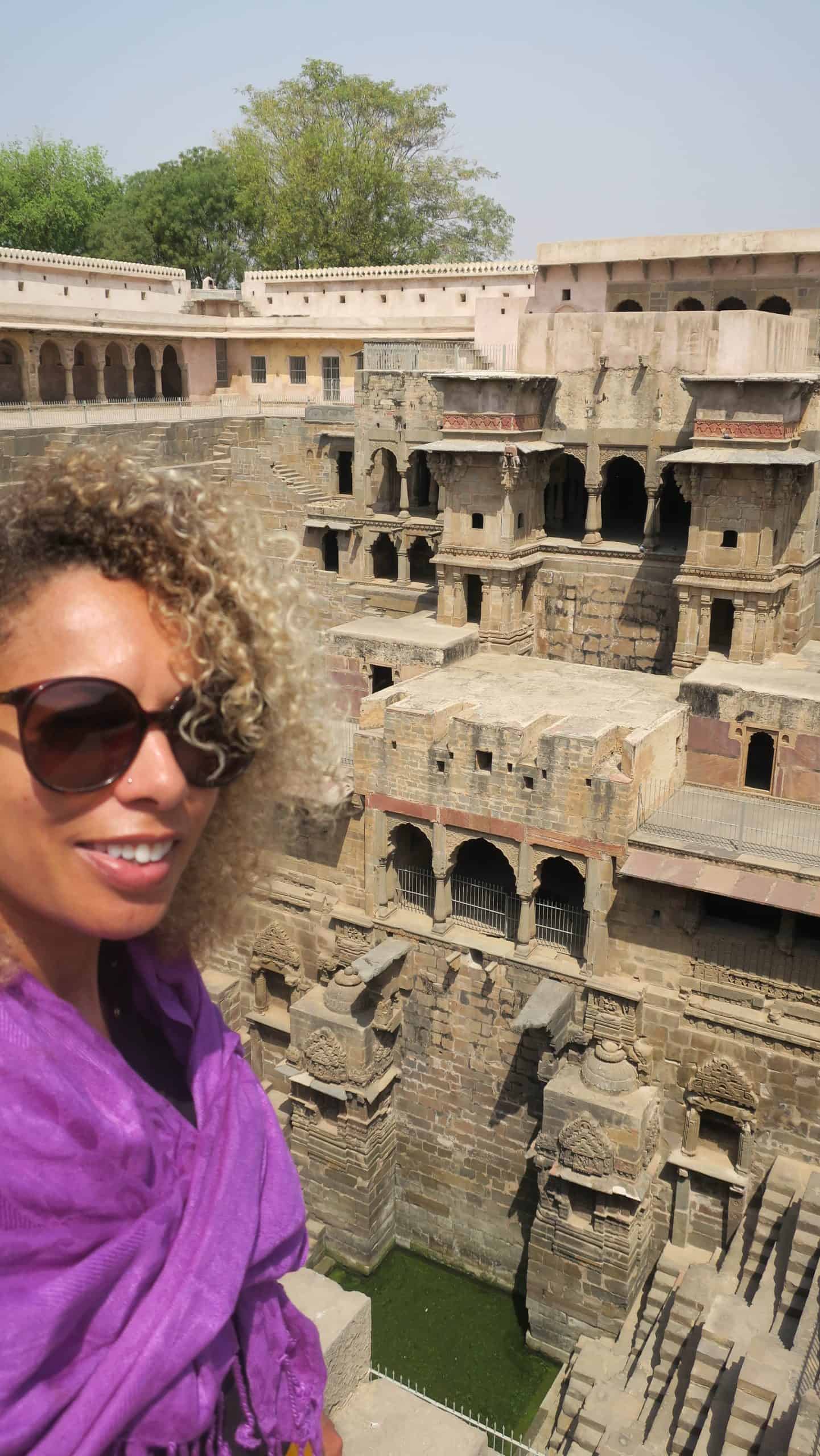 Twiggles wearing sunglasses and a purple scarf, standing beside the intricately carved stone steps of Chand Baori in Rajasthan, India.