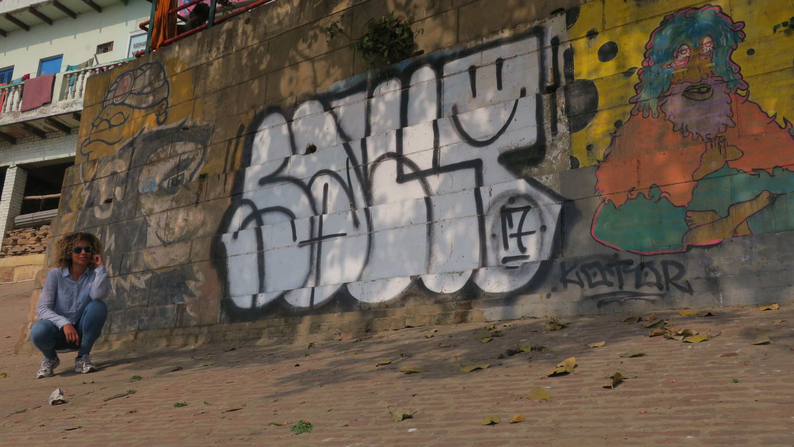 Twiggles crouching beside a colourful mural on a graffiti-covered wall in Varanasi, India.
